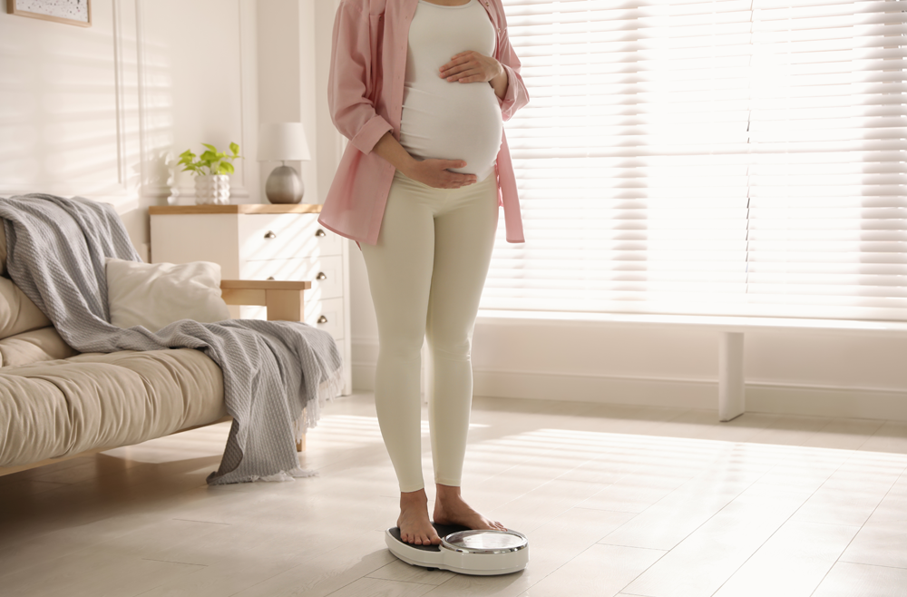A pregnant woman stands on a scale in her living room.